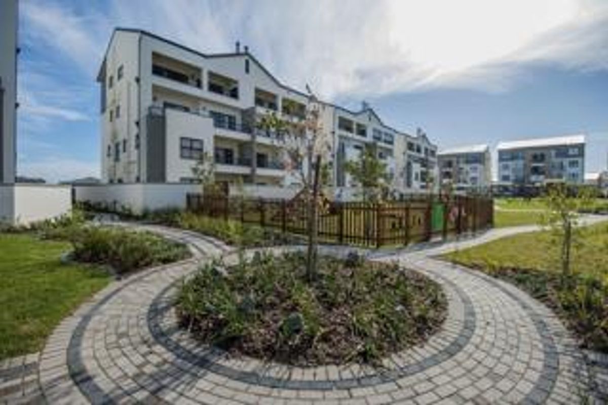 A well-maintained apartment complex exterior featuring landscaped gardens, paved walkways, and a fenced playground area for residents.