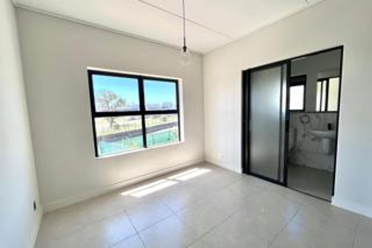 A bright, modern, empty bedroom featuring large black-framed windows, tiled flooring, and an en-suite bathroom accessible via a sliding door.