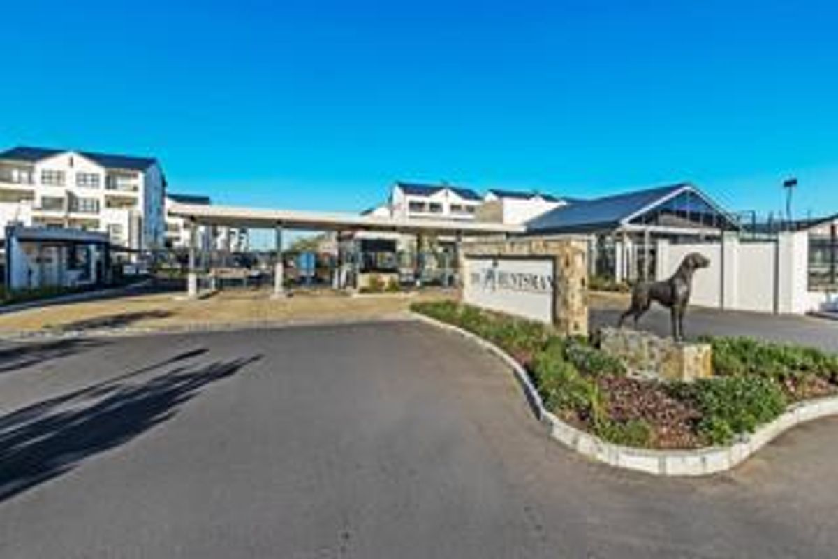 The entrance to The Huntsman apartment complex, featuring a secure gatehouse, paved driveway, and landscaped signage area.