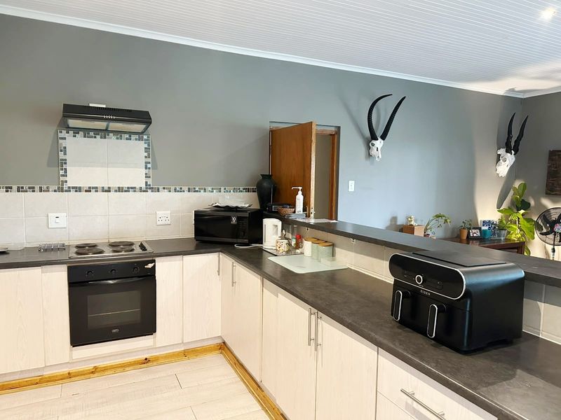 A functional kitchen featuring light-toned cabinetry, dark countertops, a built-in oven with hob, and a tiled backsplash, opening into an adjacent living area.