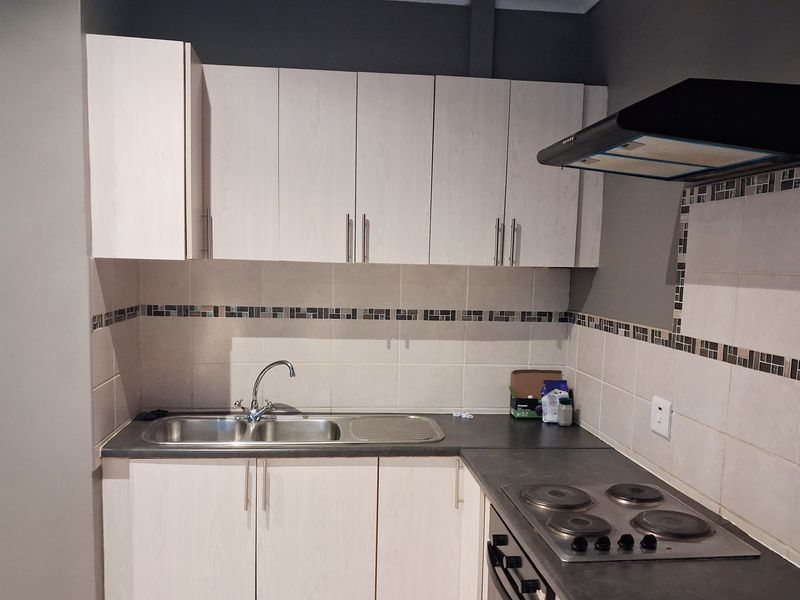A functional kitchen featuring light wood-finish cabinetry, a stainless steel sink, integrated electric hob, oven, and extractor fan with a decorative tiled splashback.