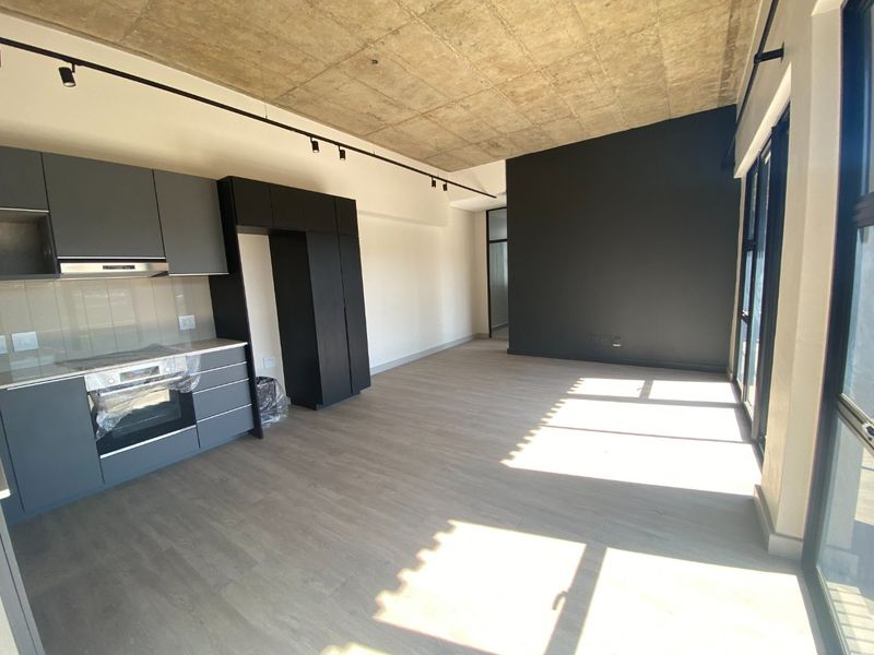 Modern open-plan living area featuring an exposed concrete ceiling, sleek dark cabinetry, integrated kitchen appliances, and large glass doors leading to an outdoor space.