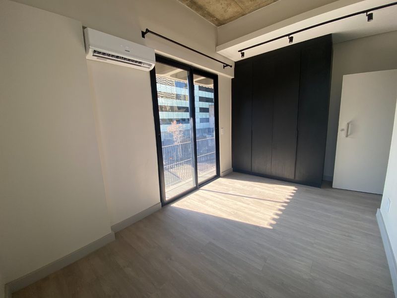 Modern bedroom featuring light wood-look flooring, a sleek black built-in wardrobe, exposed concrete ceiling, and a split-system air conditioner with balcony access.