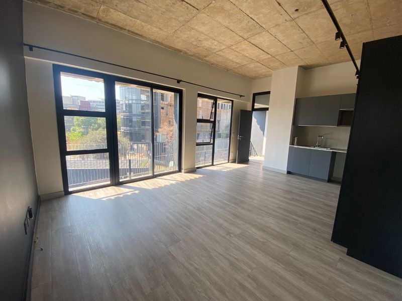 Modern open-plan apartment featuring an exposed concrete ceiling, laminate flooring, and large sliding doors leading to a balcony, with a compact integrated kitchen.