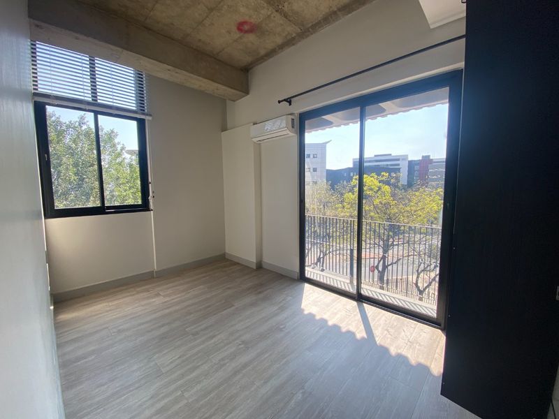 Bright bedroom featuring exposed concrete ceilings, modern laminate flooring, a split-system air conditioner, and sliding glass doors leading to a private balcony.