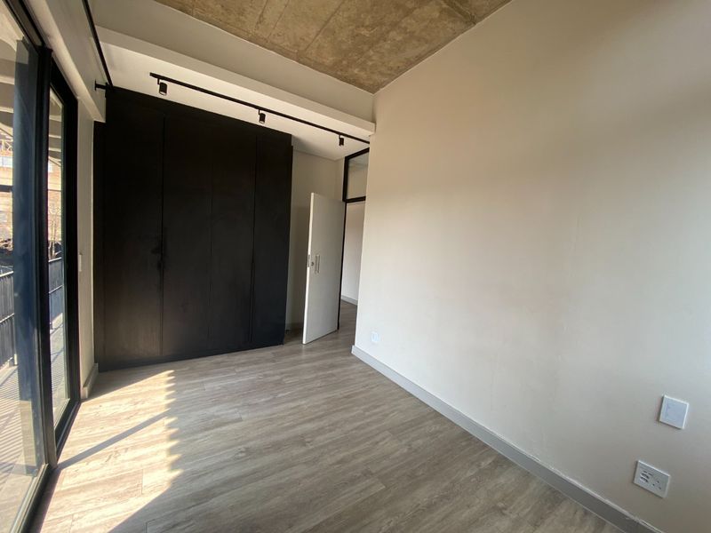 Modern bedroom featuring an exposed concrete ceiling, laminate flooring, built-in black cupboards, and a large sliding glass door leading to an outdoor area.