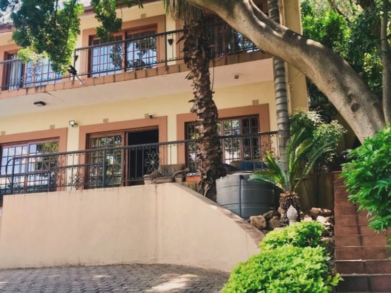 A two-story residential facade featuring wrought iron balcony railings, sliding glass doors, and a paved driveway surrounded by mature trees and landscaping.