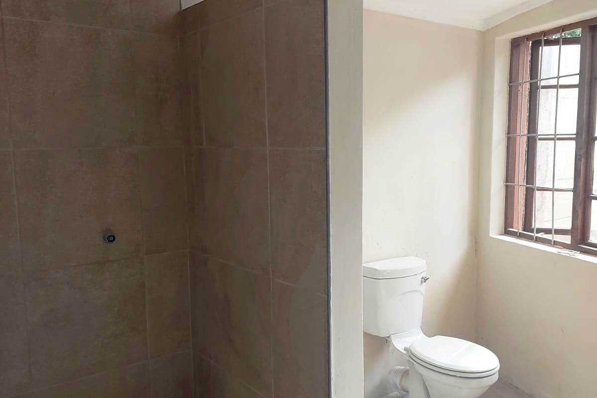 A functional bathroom featuring a tiled shower enclosure and a toilet, with natural light provided by a window with security bars.