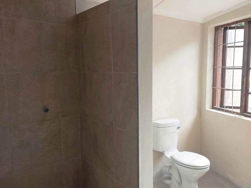 A functional bathroom featuring a tiled shower enclosure and a toilet, with natural light provided by a window with security bars.