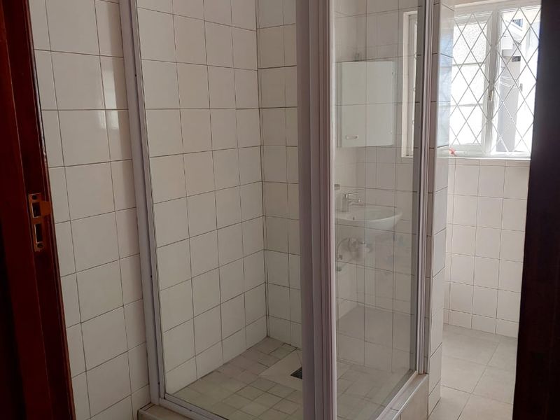 A functional bathroom featuring a tiled shower enclosure, pedestal basin, and window with security bars, presenting a clean but dated aesthetic.