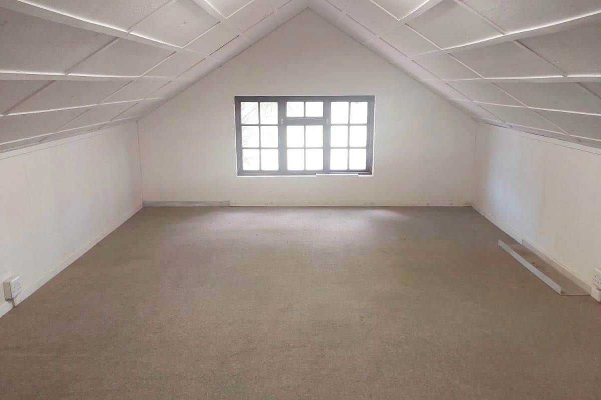 An empty attic-style bedroom featuring a pitched ceiling with exposed beams, carpeted flooring, and a multi-pane window providing natural light.