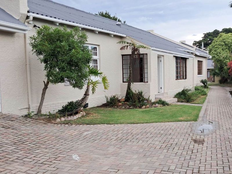 A well-maintained single-story apartment exterior featuring a pitched roof, paved driveway, and landscaped garden area with established trees.
