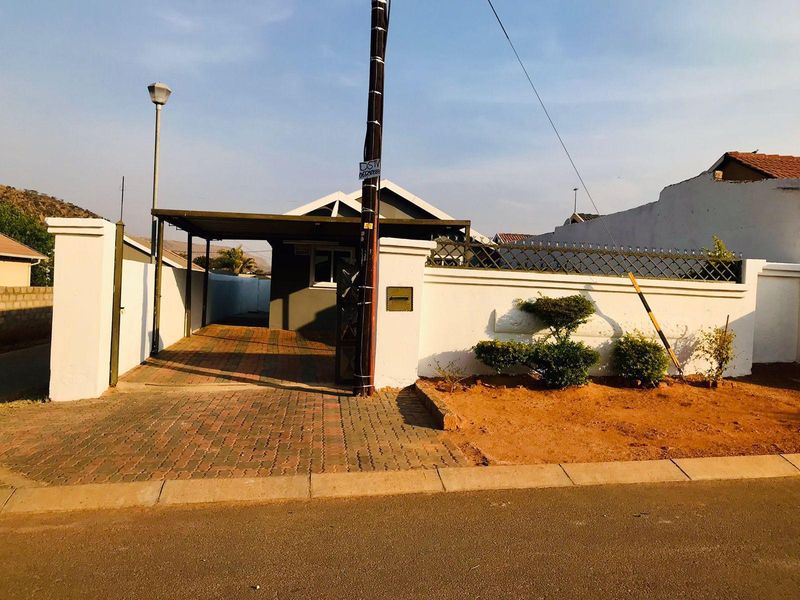 Front exterior view of the property featuring a paved driveway, covered carport, and a secure perimeter wall with integrated fencing.