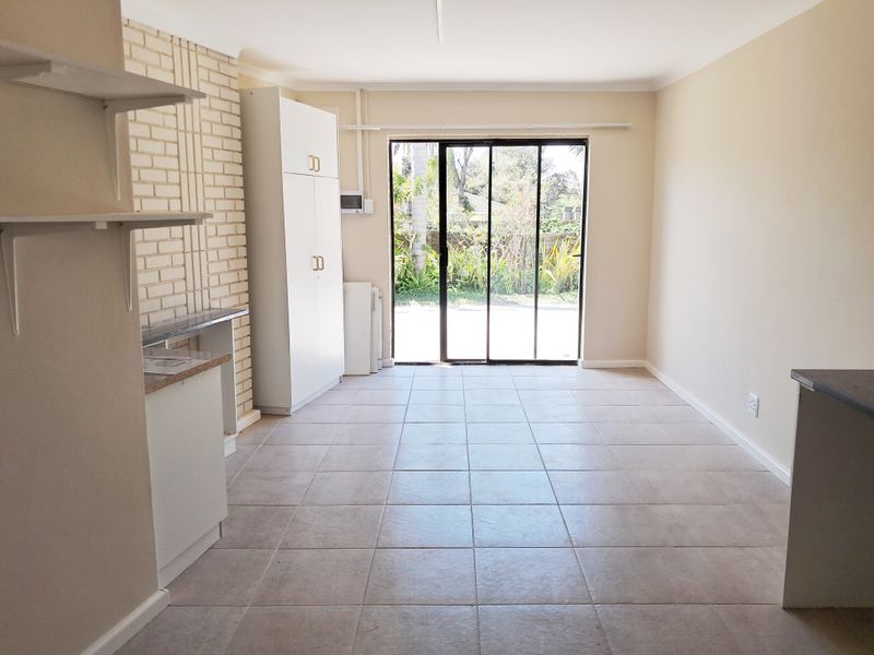 An open-plan living area featuring tiled floors, a brick feature wall, built-in storage, and a sliding glass door leading to the garden.