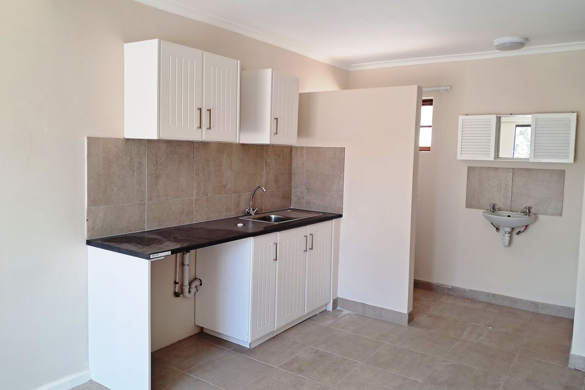 A functional kitchen area featuring white cabinetry, tiled splashbacks, a stainless steel sink, and an additional wall-mounted basin in an open-plan layout.