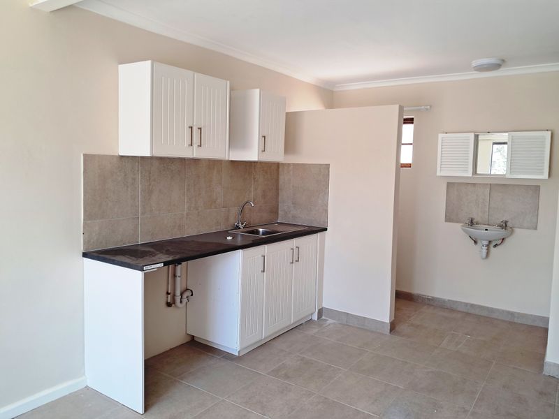 A functional kitchen area featuring white cabinetry, tiled splashbacks, a stainless steel sink, and an additional wall-mounted basin in an open-plan layout.