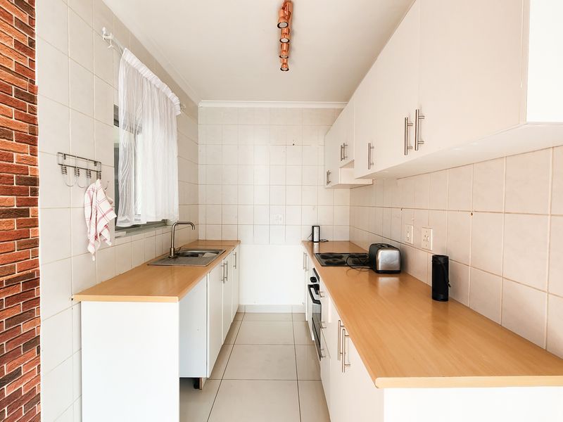 A functional galley-style kitchen featuring white cabinetry, wood-look laminate countertops, a built-in oven and hob, and a distinctive brick feature wall.