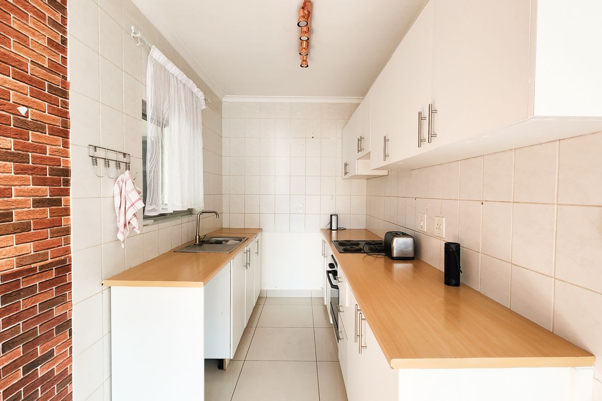 A functional galley-style kitchen featuring white cabinetry, wood-look laminate countertops, a built-in oven and hob, and a distinctive brick feature wall.