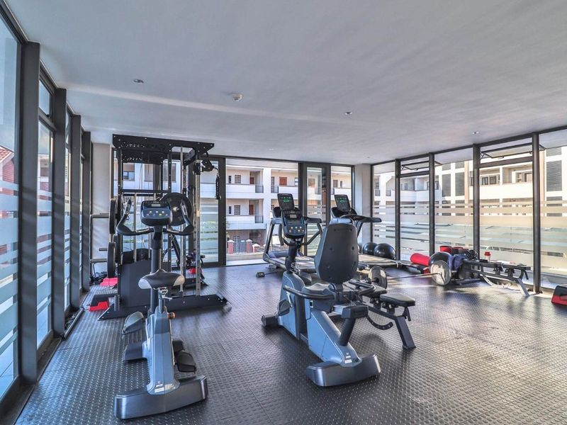 A well-lit apartment complex gym featuring rubberized flooring and floor-to-ceiling windows overlooking the surrounding residential buildings.