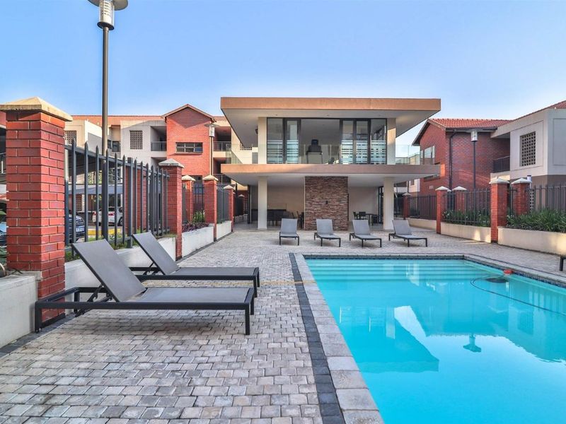 A well-maintained communal pool area featuring brick paving, a stone feature wall, and a covered patio area within a secure apartment complex.