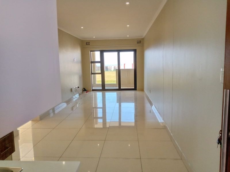 A bright, empty open-plan living area featuring polished porcelain floor tiles, recessed downlights, and a large sliding glass door leading to a balcony.