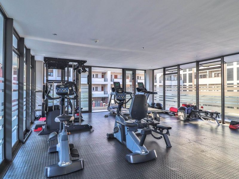 A well-lit, modern apartment gym featuring rubberized flooring and expansive floor-to-ceiling windows overlooking the complex.