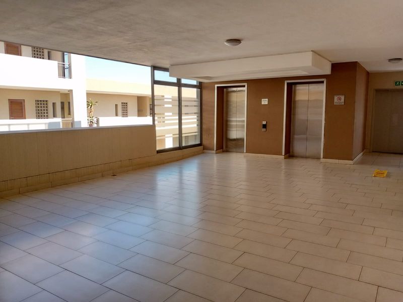 A spacious, well-maintained apartment building hallway featuring tiled flooring, dual elevators, and large windows providing natural light.
