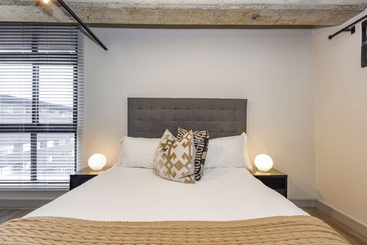 A modern bedroom featuring an exposed concrete ceiling, large window with venetian blinds, and a clean, contemporary aesthetic.