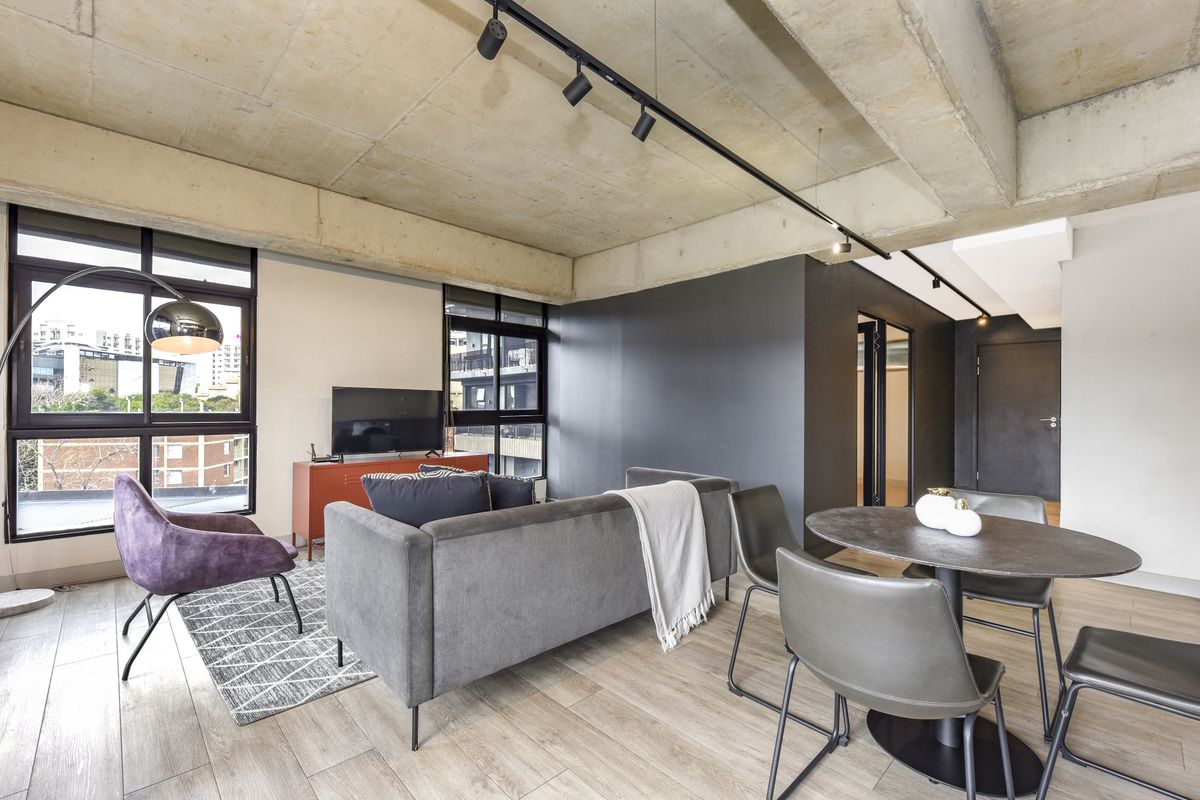 Modern open-plan living and dining area featuring an exposed concrete ceiling, track lighting, and large windows with urban views.