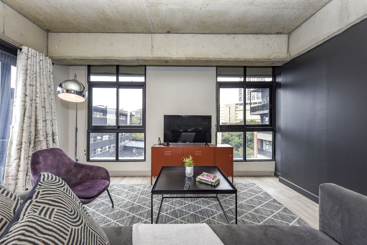 Modern living room featuring an industrial-style exposed concrete ceiling, large windows providing natural light, and contemporary laminate flooring.