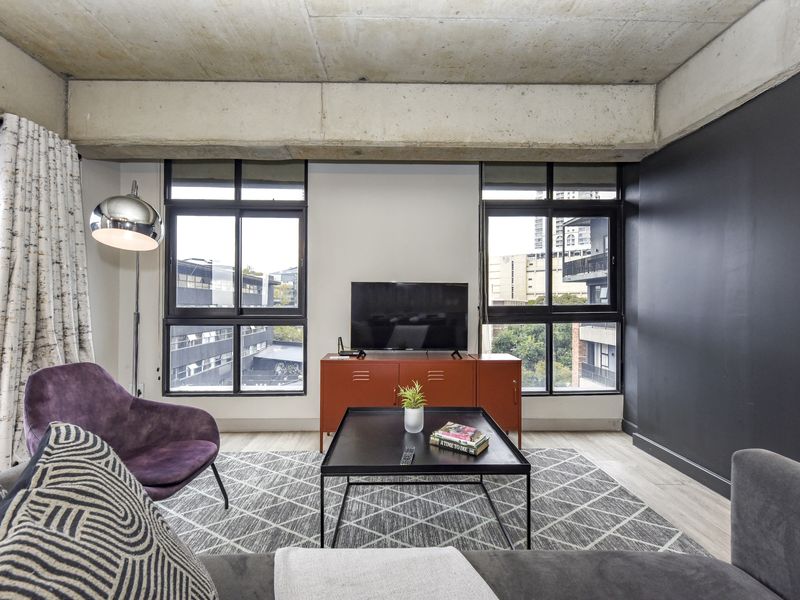 Modern living room featuring an industrial-style exposed concrete ceiling, large windows providing natural light, and contemporary laminate flooring.