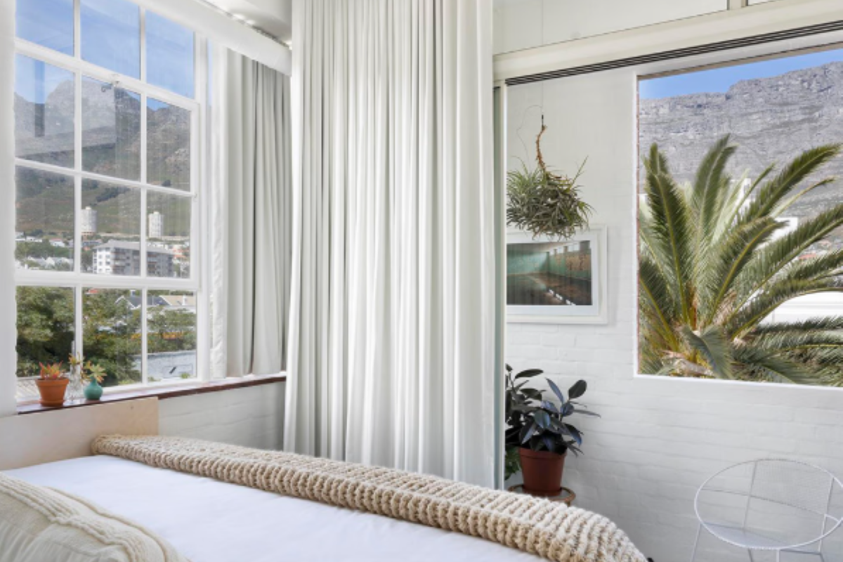 A bright bedroom featuring large windows with mountain views, white painted brick walls, and floor-to-ceiling curtains.