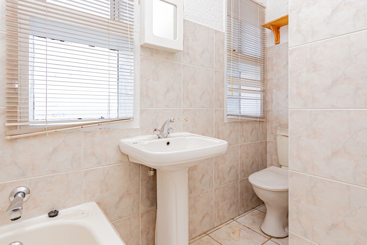 A functional bathroom featuring a pedestal basin, toilet, and bathtub with full-height wall tiling and dual windows with fitted blinds.
