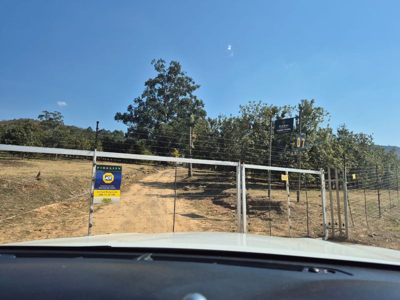 Entrance to the farm property featuring a metal gate, electric fencing, and a dirt driveway leading into the landscape.