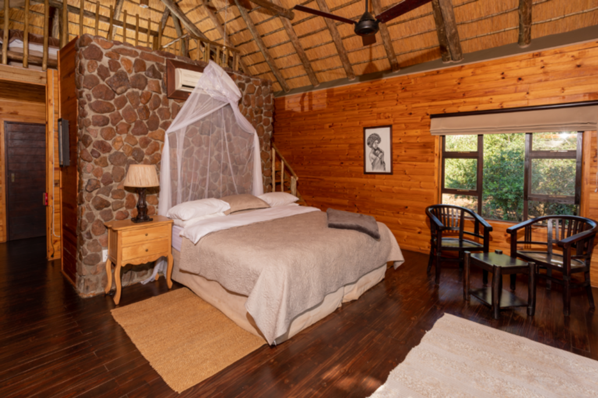 A rustic bedroom featuring a high thatched ceiling with exposed timber beams, a stone feature wall, and warm timber wall cladding.