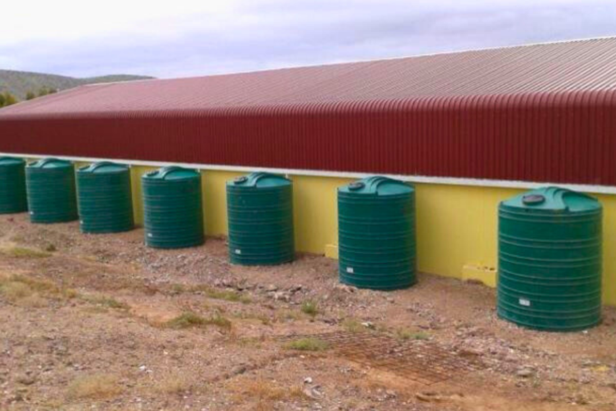 Exterior view of a farm building featuring a corrugated metal roof and a row of large green plastic water storage tanks.