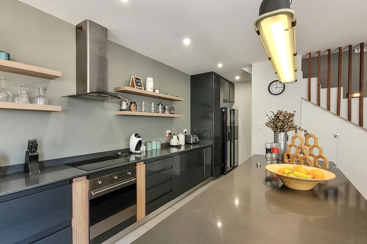 Modern kitchen featuring sleek dark cabinetry, stone countertops, built-in oven, hob, and extractor fan, complemented by floating timber shelving and an adjacent staircase.