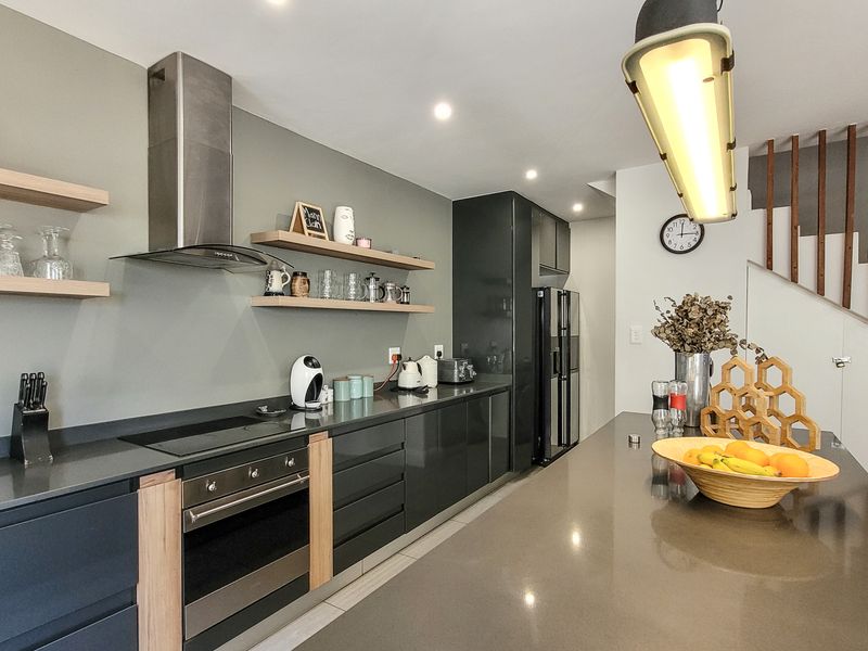 Modern kitchen featuring sleek dark cabinetry, stone countertops, built-in oven, hob, and extractor fan, complemented by floating timber shelving and an adjacent staircase.