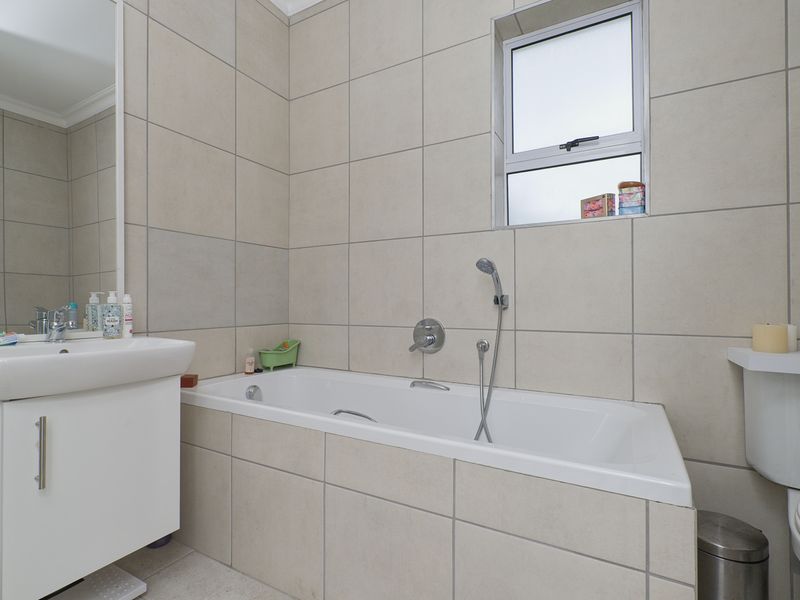 A clean, functional bathroom featuring neutral wall tiling, a built-in bathtub with a handheld shower mixer, a vanity basin, and a toilet.