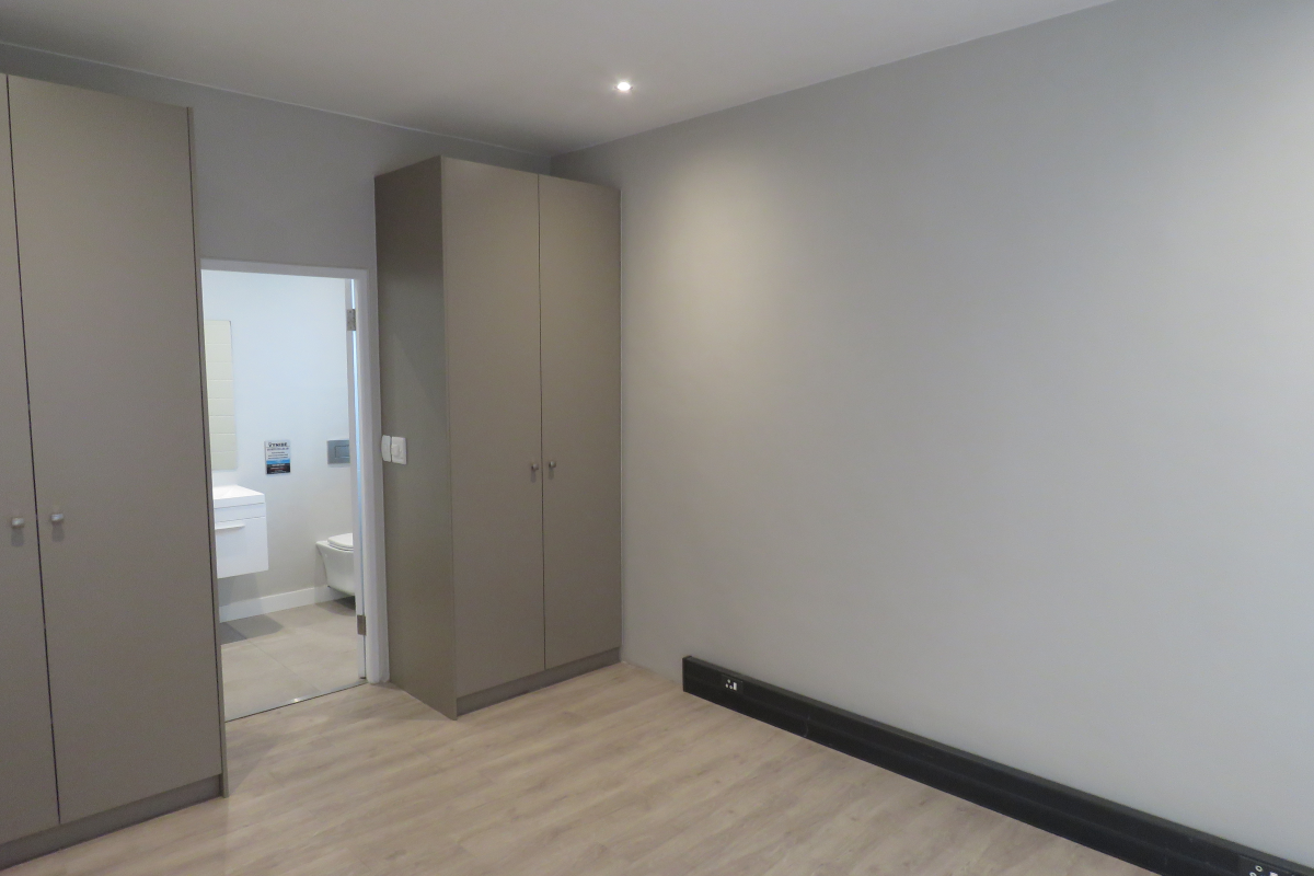 A clean, modern bedroom featuring light wood-look flooring, neutral grey walls, and built-in floor-to-ceiling cabinetry, with an adjoining en-suite bathroom.