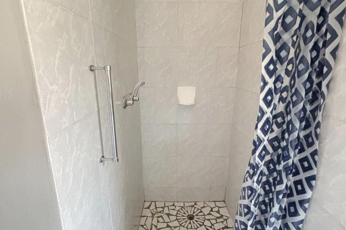 A shower enclosure featuring light-toned wall tiles and a mosaic-tiled floor, equipped with a fixed shower head and a safety grab rail.