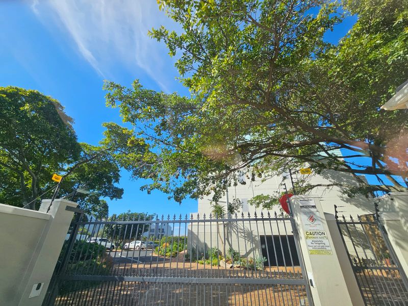 View of the apartment complex entrance featuring an automatic security gate, perimeter wall, and electric fencing under mature trees.