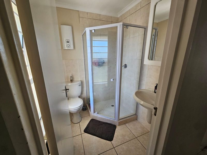 A functional bathroom featuring a corner shower enclosure, pedestal basin, toilet, and wall-mounted geyser with neutral tiled finishes throughout.