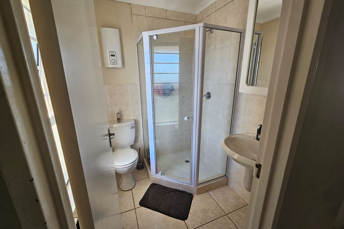 A functional bathroom featuring a corner shower enclosure, pedestal basin, toilet, and wall-mounted geyser with neutral tiled finishes throughout.