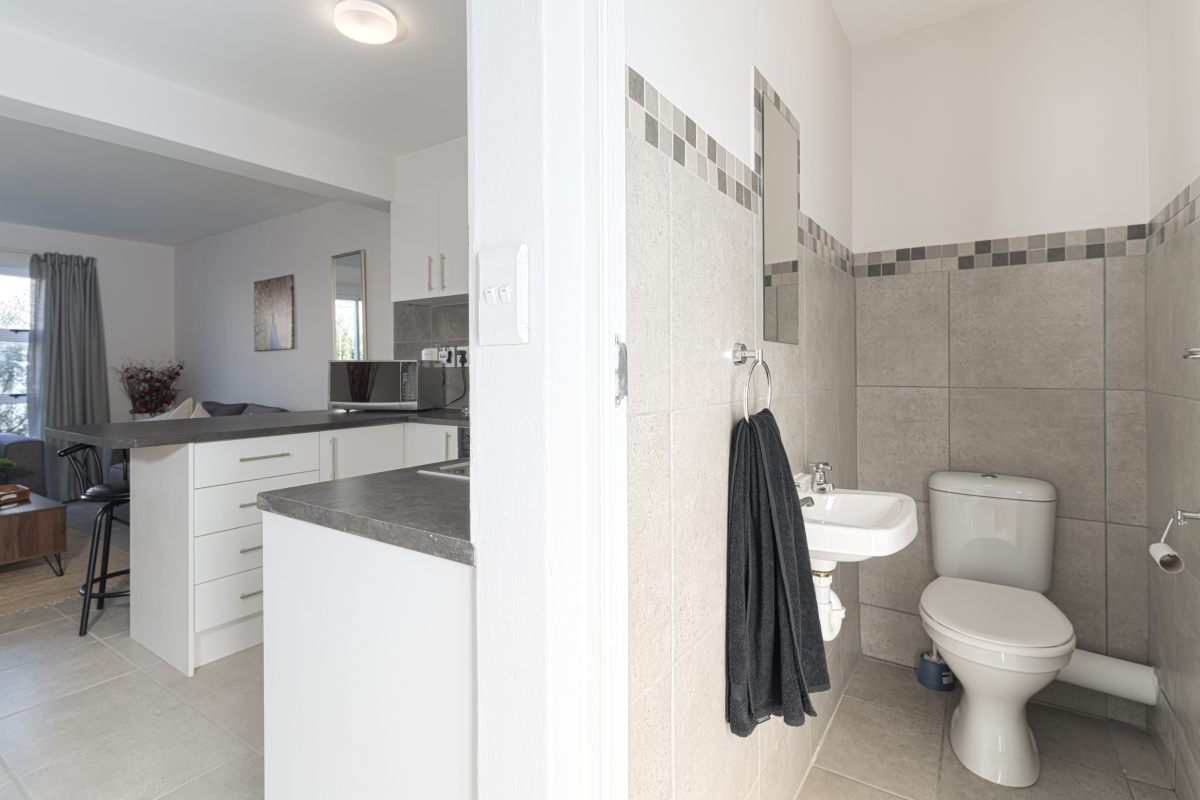 A view from a tiled bathroom featuring a wall-mounted basin and toilet, looking through to an open-plan kitchen with white cabinetry and dark countertops.
