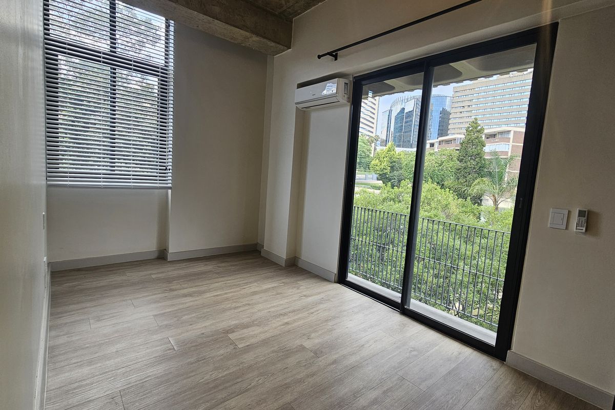 A bright, empty bedroom featuring wood-look flooring, a split-system air conditioner, and large sliding glass doors opening onto a balcony with urban views.