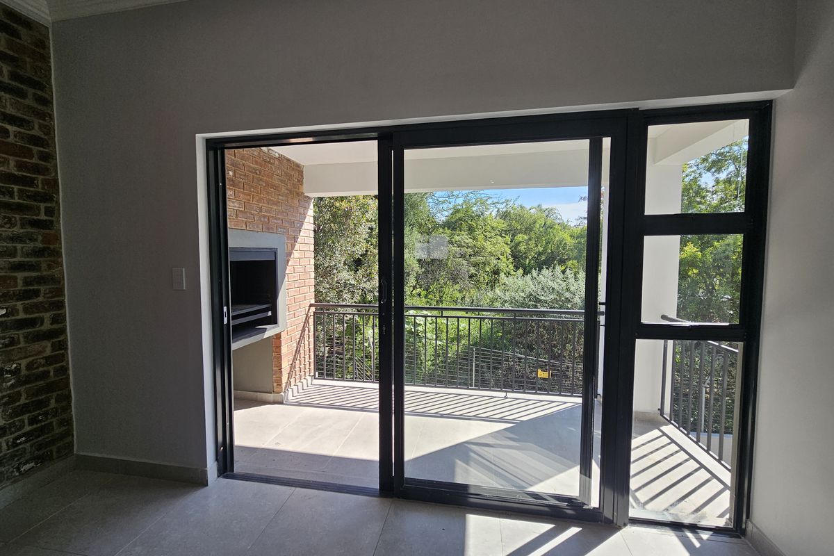 A bright living area featuring large sliding glass doors that open onto a private balcony with a built-in brick braai and leafy views.