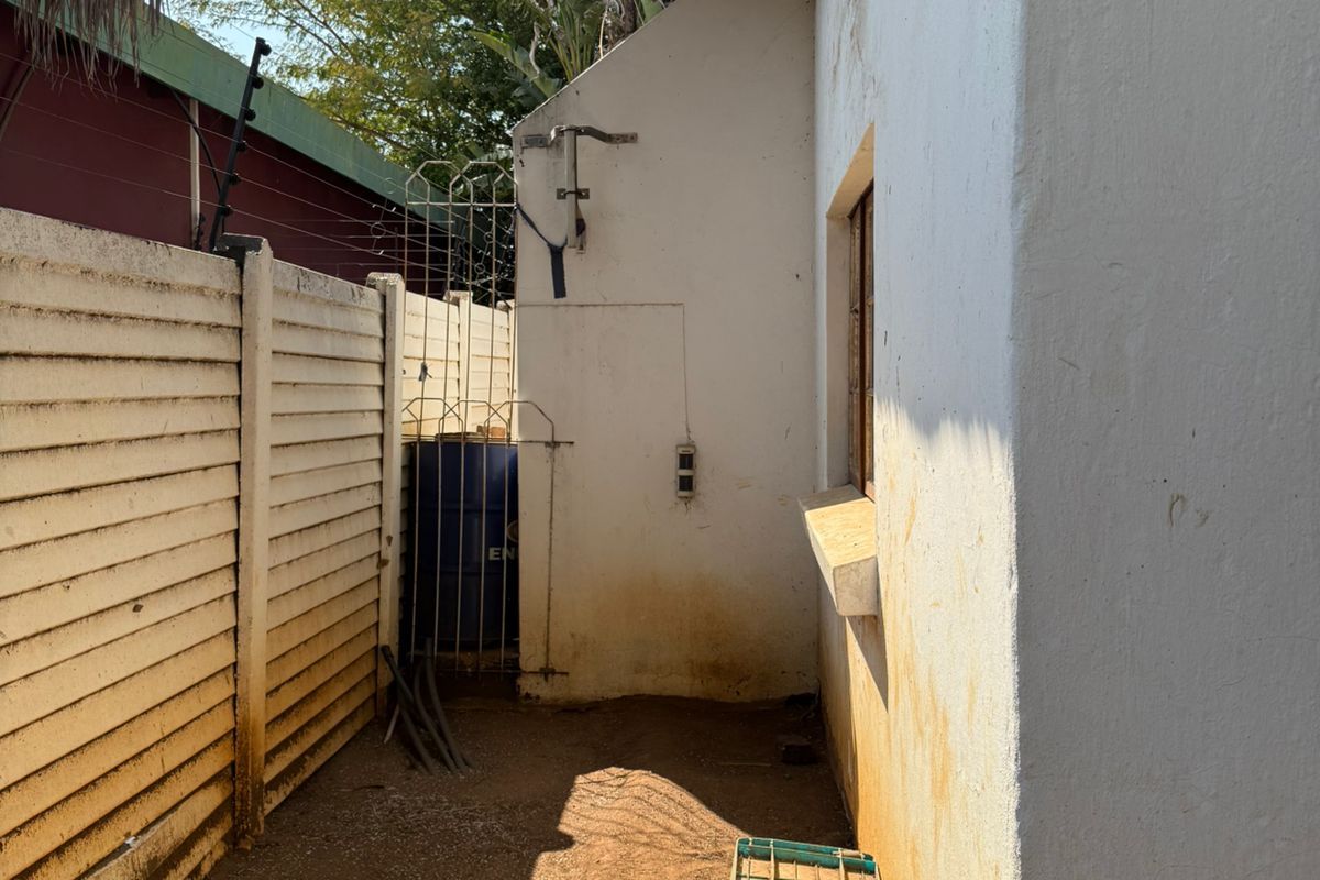 A narrow rear exterior side passage featuring a thatched roof overhang, precast concrete boundary wall with electric fencing, and weathered exterior wall finishes.