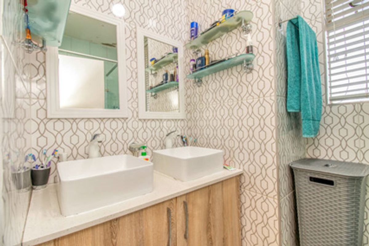 A bathroom featuring a double vanity with vessel sinks, wood-look cabinetry, patterned wall tiles, and a window with fitted blinds.