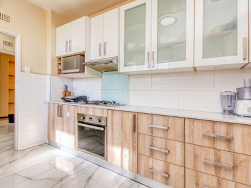 A modern kitchen featuring wood-grain cabinetry, white upper cabinets with glass inserts, a built-in oven, gas hob, extractor fan, and marble-look tiled flooring.